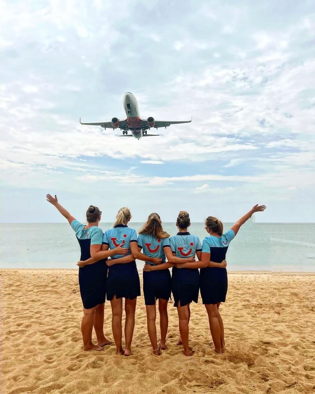 TUI Reiseleiter, Arm in Arm am Strand, winken einem Flugzeug zu, welches über sie hinweg fliegt