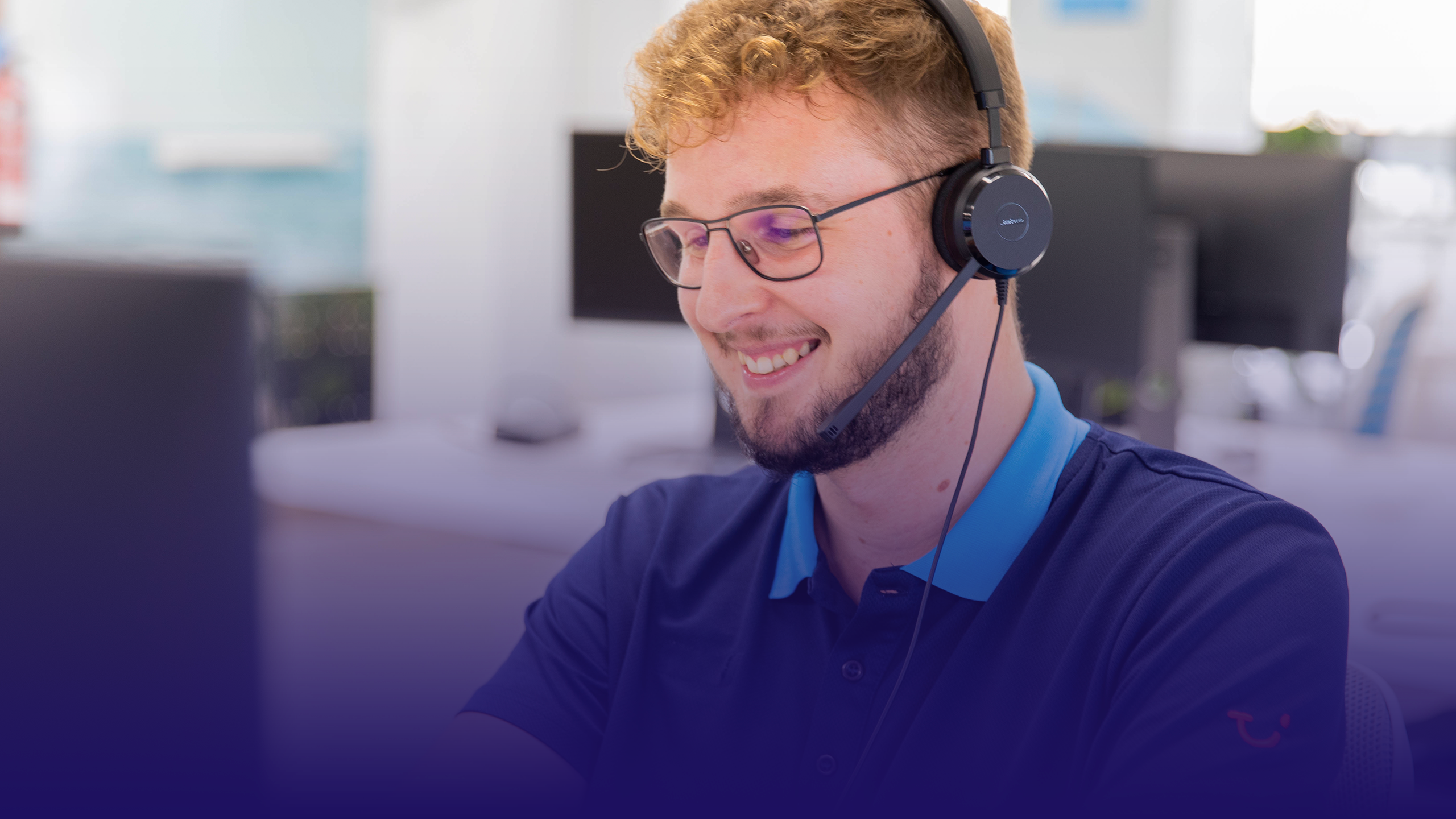 TUI Contact Center Agent lächelt im Büro seinem Computer zu und trägt ein Headset