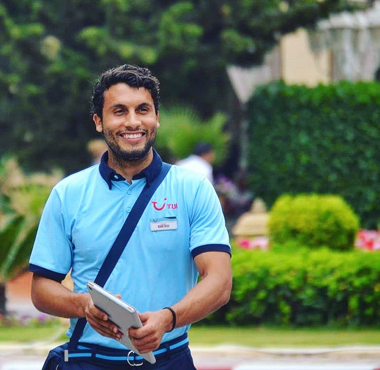 A rep smiling wearing uniform with a bag strap across their shoulder. There are green shrubs in the background.